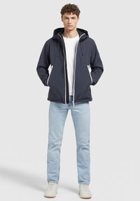 Veste coupe-vent marine avec capuche, zip blanc contrastant, poches latérales et cordons de serrage ajustables. Associée à un jean bleu clair.