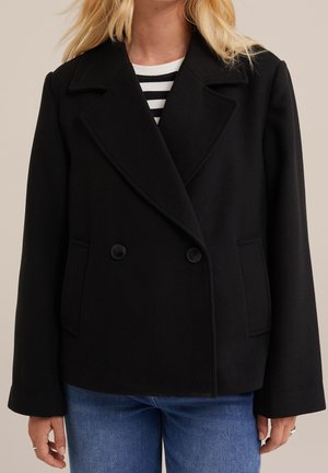 Kvinde iført en sort dobbeltradet frakke over en sort-hvid stribet top og blå jeans med blondt hår synligt.