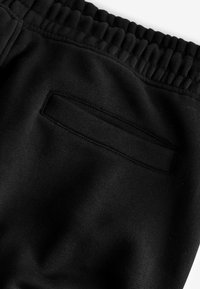 Schwarze Jogginghosen aus weichem Stoff, mit einem elastischen Bund und einer rechteckigen Tasche auf der Rückseite. Glatte Textur und minimalistisches Design.