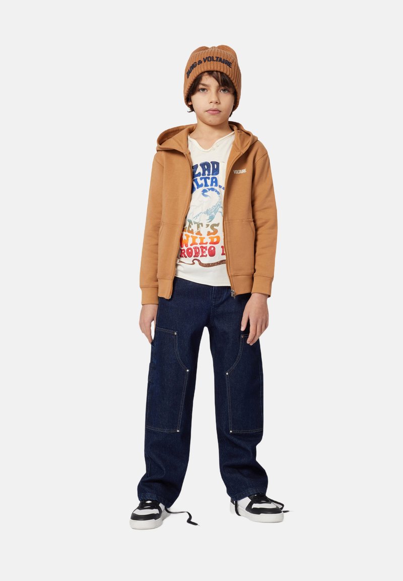 Veste à capuche marron sur un T-shirt graphique, pantalon cargo en denim bleu avec de grandes poches avant, et baskets noir et blanches. Bonnet en tricot marron.