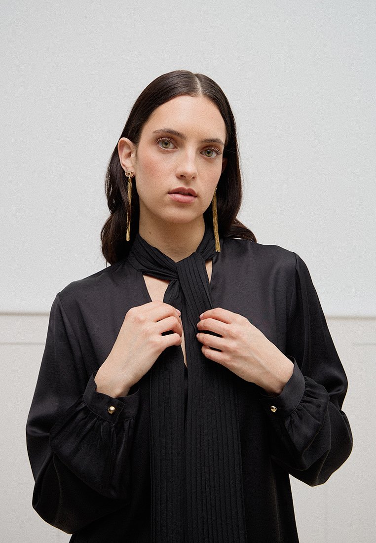 Blusa nera a maniche lunghe con dettaglio al collo plissettato e annodato, e rifiniture con bottoni sui polsini. Tessuto liscio con un design fluido.