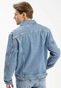 Jasnoniebieska kurtka dżinsowa o lekko wyblakłej fakturze, z kołnierzykiem, guzikami przy mankietach oraz detalami na plecach.
