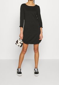 Černé šaty do délky kolen s třičtvrtinovými rukávy, s okrajem ve tvaru vlny, zkombinované s mini kabelkou s vzorem kravské srsti a černými teniskami.