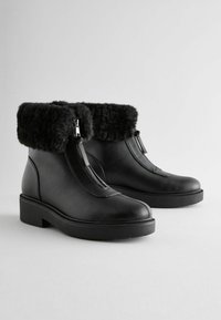 Black leather ankle boots feature a faux fur cuff, zip closure, and a thick, textured sole. Smooth surface with minimal decorative elements.