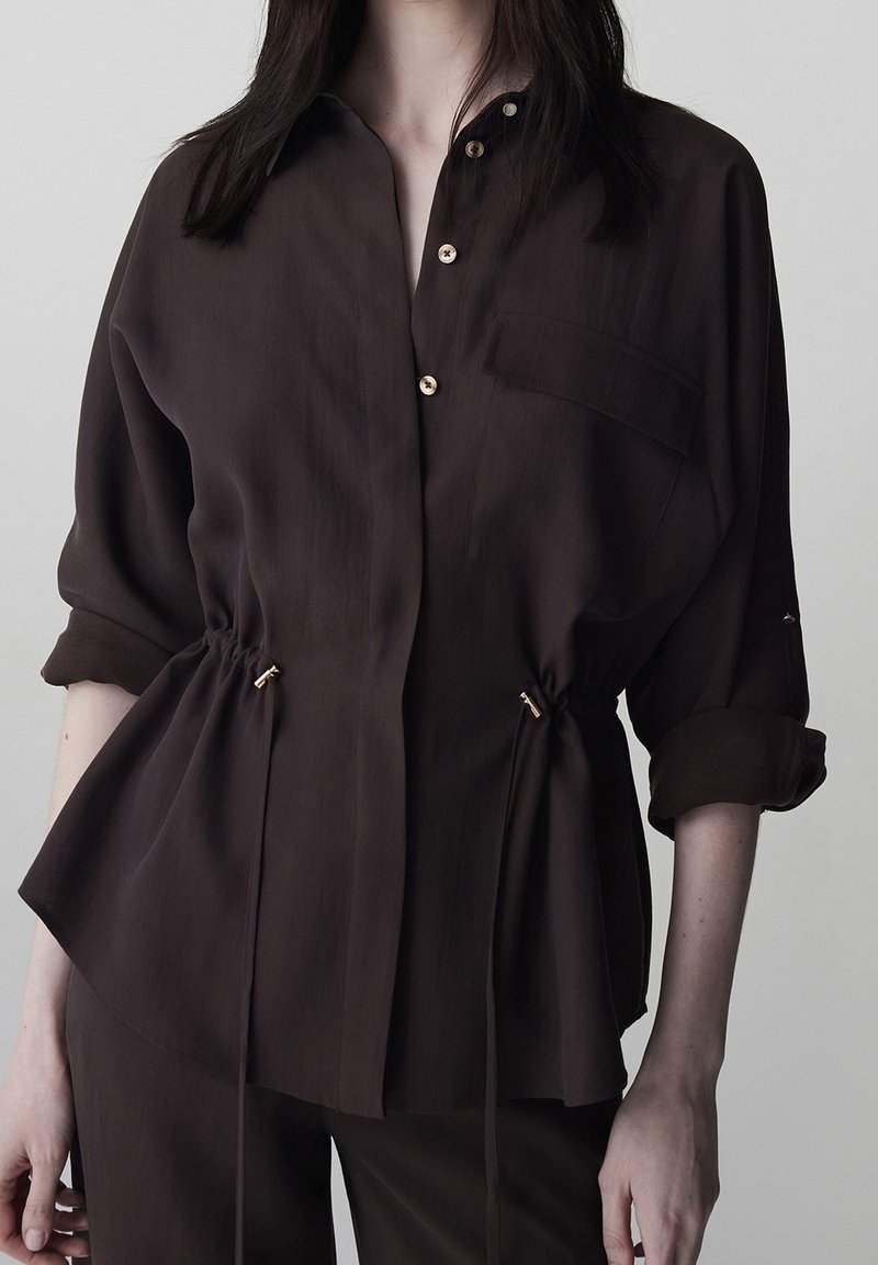 Femme portant un chemisier marron foncé à manches longues avec manches retroussées et liens ajustables à la taille, associé à un pantalon foncé assorti.
