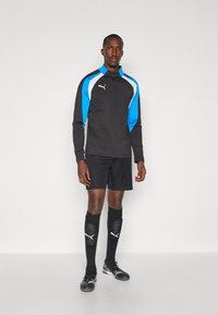 Puma treniruočių apranga, kurią sudaro juodas ilgarankovis marškinėlis su mėlynomis ir baltomis akcentais, juodos šortai ir juodos iki kelių kojinės su logotipu.