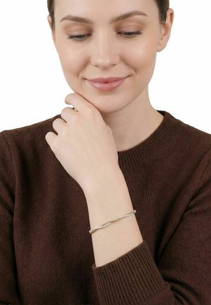 Donna con un maglione marrone che guarda verso il basso sorridendo, indossando un delicato braccialetto intrecciato in oro e argento al polso sinistro.
