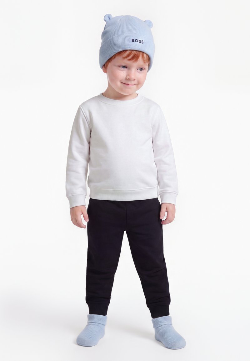 Jeune enfant portant un chapeau bleu clair avec des oreilles et le texte "BOSS", une chemise blanche à manches longues, un pantalon noir et des chaussettes bleu clair assorties.