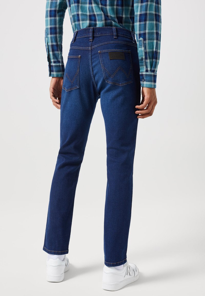 Niebieskie dżinsy o prostym kroju, z dwiema tylnymi kieszeniami i charakterystycznym wzorem przeszycia. Noszone z białymi trampkami.