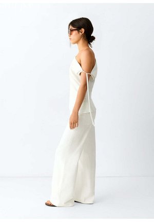 Donna che indossa un top bianco con spalle scoperte e dettaglio annodato e pantaloni bianchi a gamba larga, in piedi di profilo su uno sfondo chiaro e semplice.