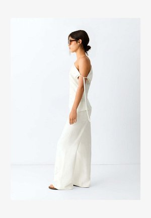 Vrouw draagt een witte off-shoulder top met strikdetail en een wijde witte broek, staand in profiel tegen een effen lichte achtergrond.