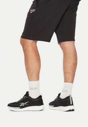 Melnas sporta kurpes ar teksturētu virsmu, baltu zoli un Reebok logotipu. Apvienotas ar baltām zeķēm un melniem šortiem, parādot kāju detaļas.