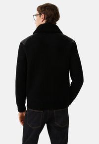 Zwarte geribbelde trui met een hoge, pluizige kraag en leren schouderaccenten, voorzien van lange mouwen en een nauwsluitende silhout.