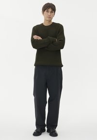 Jauns vīrietis stāv ar rokām krustā, ģērbies teksturētā tumši zaļā džemperī, brīvās melnās biksēs un melnos lace-up apavos uz balta fona.