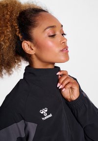 Svart jacka av lättviktsmaterial, med hög krage, grå detaljer och en vit logotyp på bröstet. Slät textur.