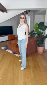 Top bianco senza maniche, jeans svasati azzurri chiari e scarpe bianche. Gli accessori includono occhiali da sole marroni e una collana. Divano angolare marrone sullo sfondo.