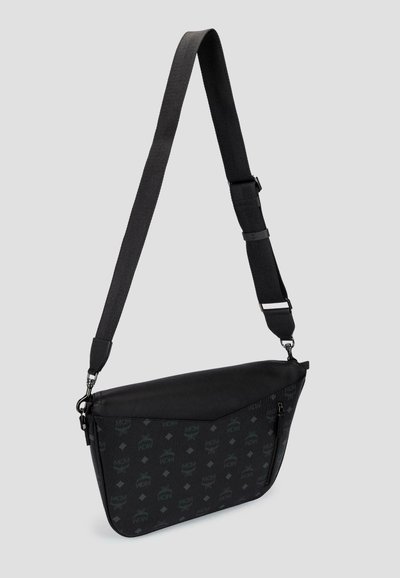 Zwarte crossbodytas met verstelbare riem, MCM-logo patroon en een voorvak met rits, afgebeeld tegen een effen lichtgrijze achtergrond.