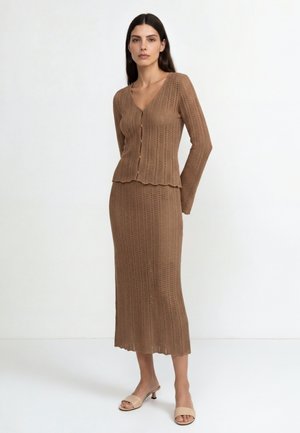 Femme portant un cardigan marron en maille boutonné et une longue jupe assortie avec des sandales beiges à talons ouverts, debout devant un fond uni.