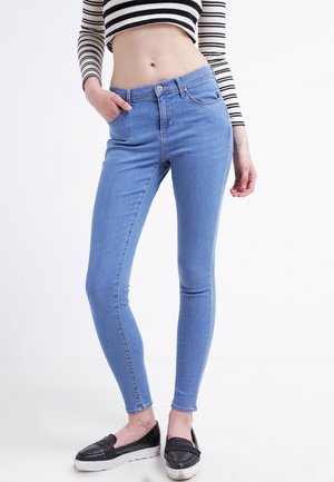 Femme portant un jean skinny bleu clair, un haut court à manches longues rayé noir et blanc, et des chaussures noires sans lacets, debout devant un fond blanc.