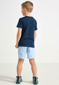 Marinblå T-shirt med kort ärm, ljusblå shorts med segelbåtsmönster och mönstrade blå sneakers med gröna sulor.