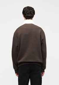 Καφέ φούτερ με λευκό γιακά και πλευρικές λεπτομέρειες, με άνετη εφαρμογή και υφή υφάσματος. Φαίνεται από πίσω, χωρίς ορατά λογότυπα.