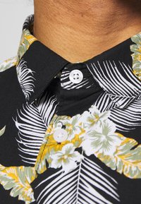 Zwarte shirt met een bloemenpatroon met witte, groene en gele bladeren. Kraagontwerp met twee witte knopen bij de nek.