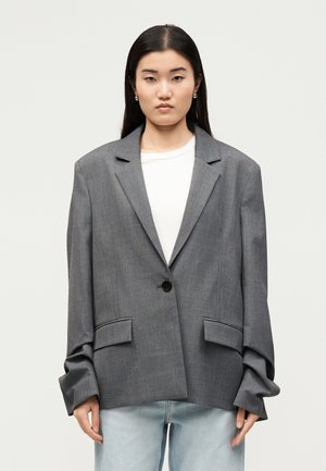 Jeune femme aux longs cheveux foncés portant un blazer gris oversize, une chemise blanche et un jean bleu clair, debout devant un fond clair uni.