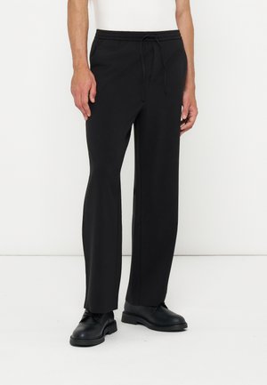 Homme portant un pantalon noir droit avec taille à cordon et des chaussures noires à lacets, debout devant un fond blanc.