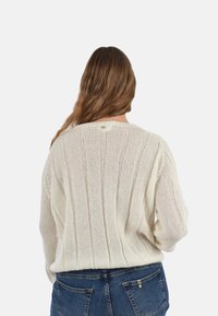 Maglione lavorato a maglia di colore crema chiaro con motivo a coste verticali, scollatura tonda e piccolo bottone decorativo sulla parte posteriore. Abbinato a jeans di denim.