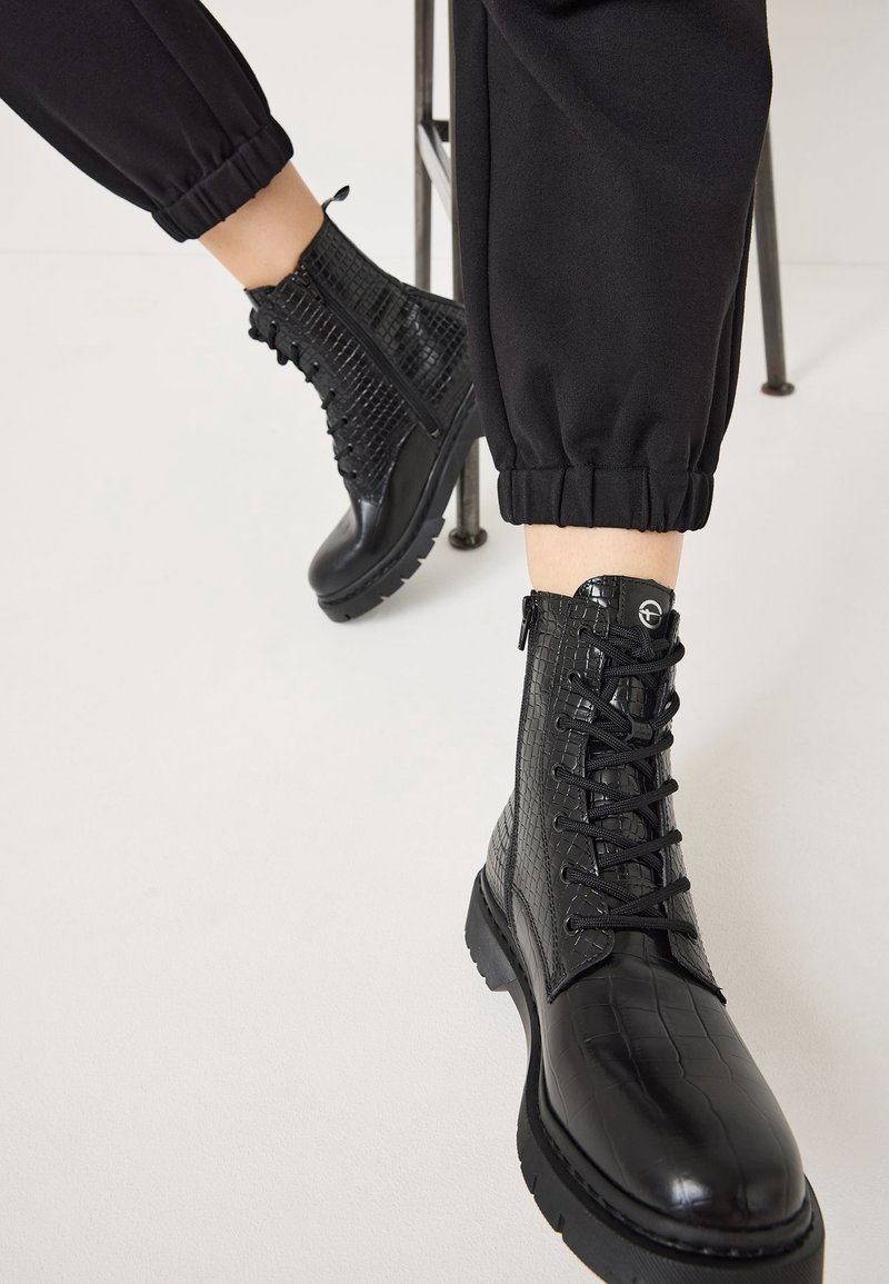Bottes courtes à lacets en cuir noir texturé, portées avec un pantalon noir retroussé, personne assise sur une chaise en métal avec un pied en avant.