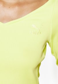 Jasnożółta, prążkowana bluzka z dekoltem w kształcie litery V i krótkimi rękawami, z białym haftowanym logo Puma na piersi.