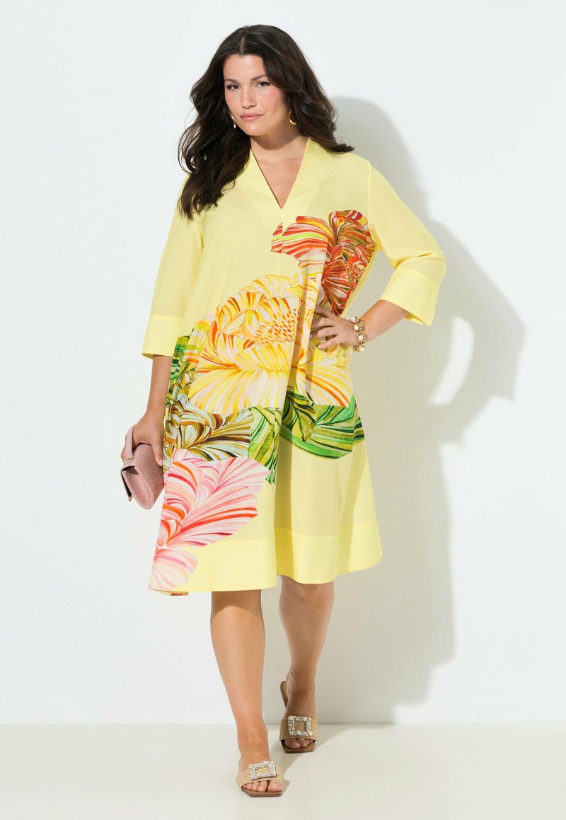 Donna con un vestito giallo a grandi stampe floreali, che tiene una pochette rosa, indossa sandali e accessori dorati, in piedi davanti a uno sfondo bianco.