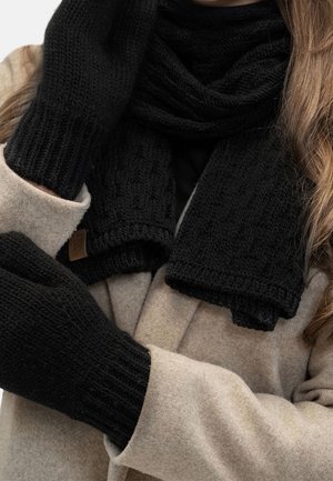 Personne portant un manteau beige, une écharpe en tricot noire et des moufles en tricot noires, avec des cheveux bruns lâches visibles au-dessus du manteau.