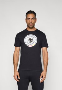 Fekete pamut póló, kerek logóval, amelyen egy sas és a "Deutscher Fussball-Bund" felirat látható, kiemelő színek piros, sárga és fekete.