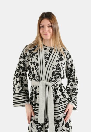 Giovane donna che indossa un cardigan con cintura e fantasia e pantaloni coordinati con motivi floreali e a righe nere su sfondo grigio chiaro.