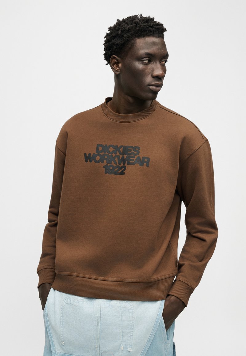 Man wearing a brown sweatshirt with "Dickies Workwear 1922" text, light blue pants, hands in pockets, looking to the side on a plain background.