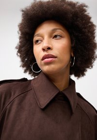 Cappotto in suede marrone con un ampio colletto e dettagli sulle spalle. La modella ha capelli ricci e indossa orecchini a cerchio argentati.