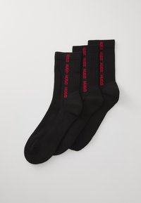 Trois paires de chaussettes noires de type crew avec un logo rouge "HUGO" vertical le long des côtés, posées à plat sur un fond blanc.