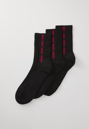 Drei Paar schwarze Crew-Socken mit rotem vertikalem "HUGO"-Branding an den Seiten, flach auf einem weißen Hintergrund ausgelegt.