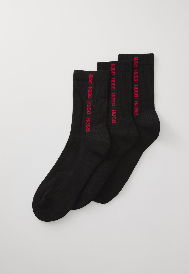 Trois paires de chaussettes noires de type crew avec un logo rouge "HUGO" vertical le long des côtés, posées à plat sur un fond blanc.