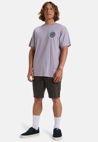 Camiseta de algodón lavanda con un logo circular en el pecho, combinada con pantalones cortos gris oscuro, calcetines blancos y zapatillas azul marino.