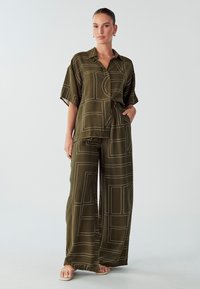 Olivgrünes, kurzärmeliges Hemd mit Knopfleiste und einem Gürtel in der Taille, kombiniert mit weit geschnittenen Hosen mit geometrischen weißen Mustern.