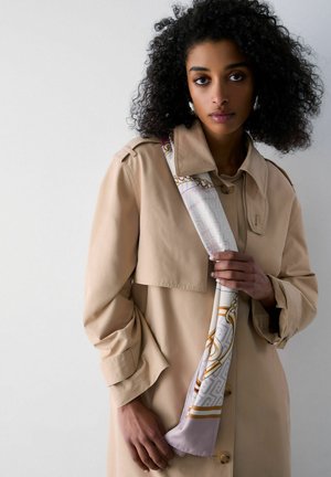 Woman with curly hair wearing a beige trench coat, holding a patterned silk scarf draped over her shoulder against a plain background.