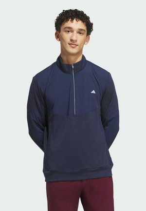 Junger Mann mit lockigem Haar, der einen marineblauen Adidas-Pullover mit halbem Reißverschluss und burgunderfarbene Hose trägt, steht mit den Händen im Rücken.