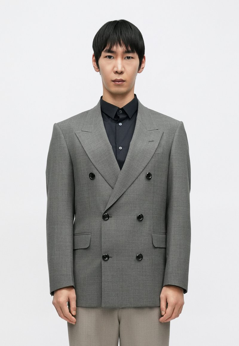 Dwurzędowa szara marynarka z teksturowanej tkaniny, z czarnymi guzikami, ostrymi klapami i patkowymi kieszeniami, noszona na czarnej koszuli.