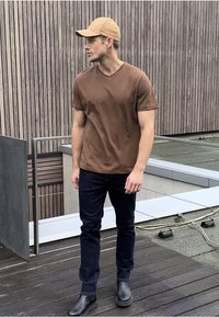 Camiseta de algodón marrón, jeans oscuros y botas de cuero negras. La persona lleva una gorra de béisbol marrón clara y está de pie sobre una superficie de madera.