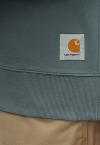 Γκρι ριμπ ύφους φούτερ με λευκή ετικέτα που φέρει πορτοκαλί λογότυπο Carhartt. Απαλή υφή, ορατές ραφές και άνετη εφαρμογή.