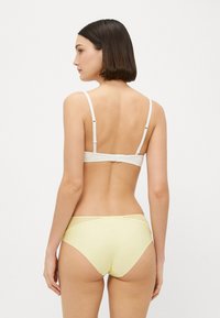 Fehér bralette vékony pántokkal és kapoccsal, sárga bikini alsóval párosítva, amelynek derékrészén texturált hálós betét található.