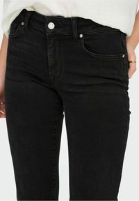 Primer plano de una persona que lleva jeans de mezclilla negros con botones y remaches plateados, combinados con una camisa blanca metida por dentro.