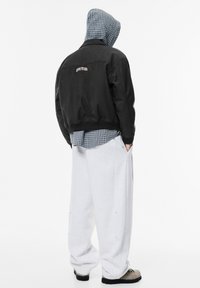 Musta bomber-takki, jossa logo takana, sinisen ruudullisen paidan päällä. Löysät harmaat collegehousut ja rakenteelliset harmaat jalkineet.